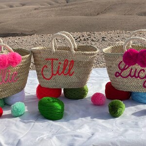 May include: Three straw tote bags with personalized names, Ellie, Jill, and Lucy, decorated with pink pom poms. The bags are sitting on a white surface with colorful pom poms.