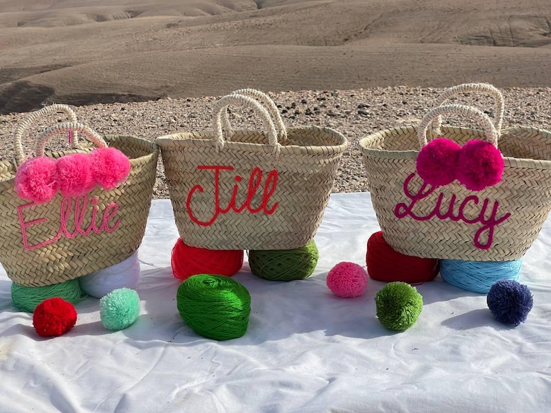 May include: Three straw tote bags with personalized names, Ellie, Jill, and Lucy, decorated with pink pom poms. The bags are sitting on a white surface with colorful pom poms.