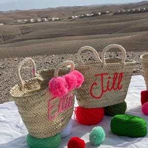 May include: Two straw tote bags with pom poms and personalized embroidery. The bags are labeled "Jill" and "Ellie". The bags are surrounded by colorful yarn balls and pom poms.