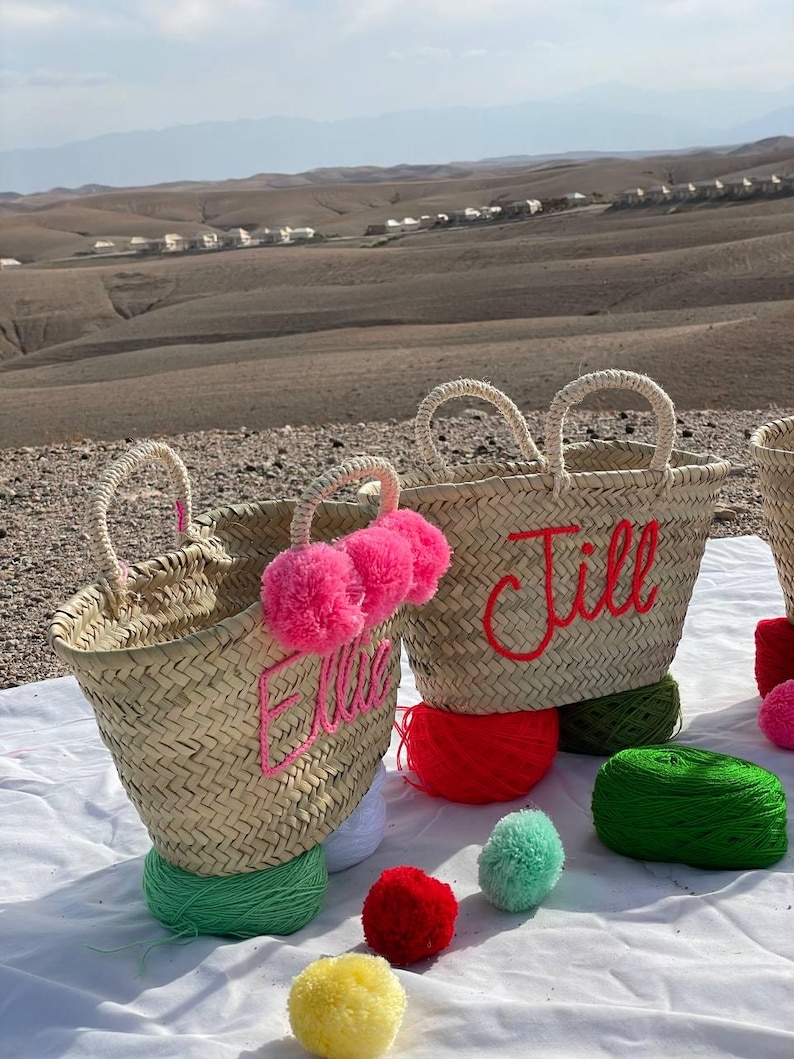 May include: Two straw tote bags with pom poms and personalized embroidery. The bags are labeled "Jill" and "Ellie". The bags are surrounded by colorful yarn balls and pom poms.