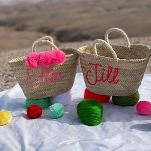 May include: Two straw tote bags with pom poms and embroidered names "Ellie" and "Jill" in red thread. The bags are sitting on a white blanket with colorful yarn balls.