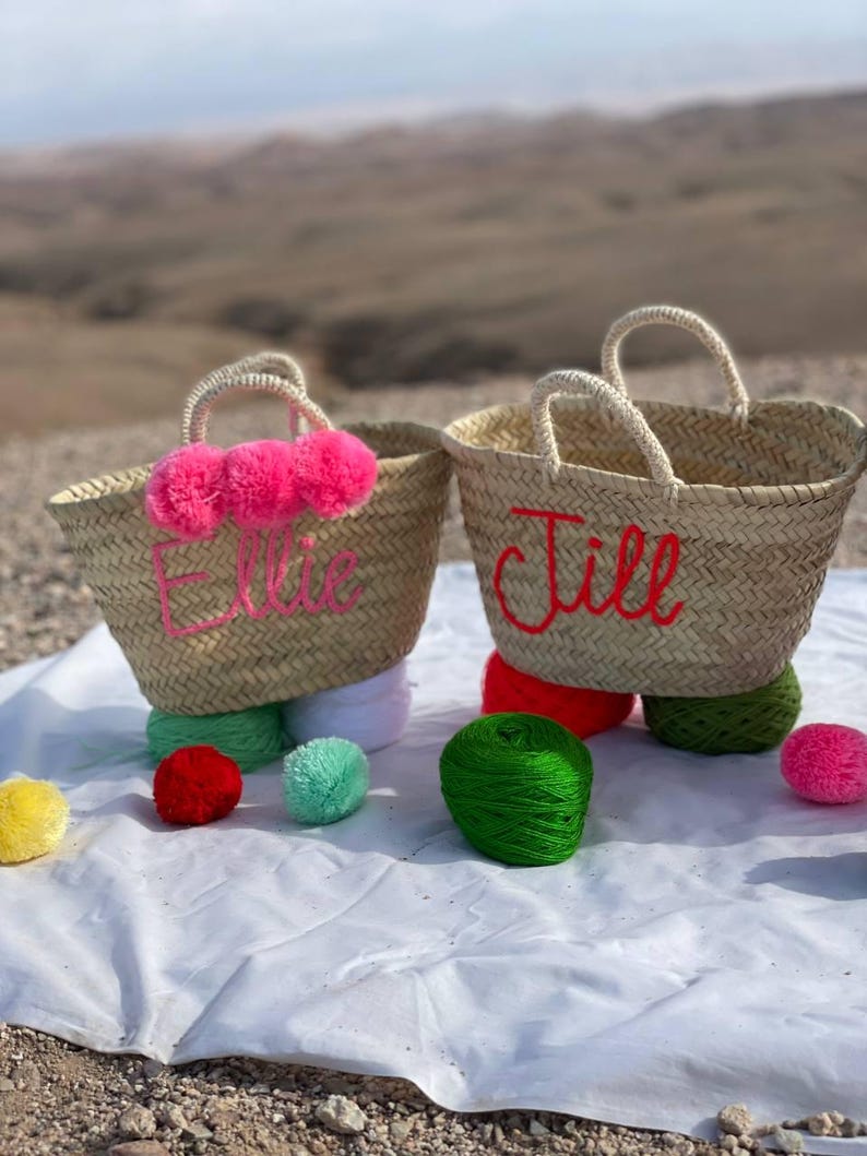 May include: Two straw tote bags with pom poms and embroidered names "Ellie" and "Jill" in red thread. The bags are sitting on a white blanket with colorful yarn balls.