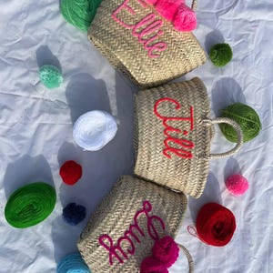 May include: Three straw tote bags with pom poms and personalized names. The bags are Ella,  Tila, and Roma. The bags are surrounded by colorful yarn balls.