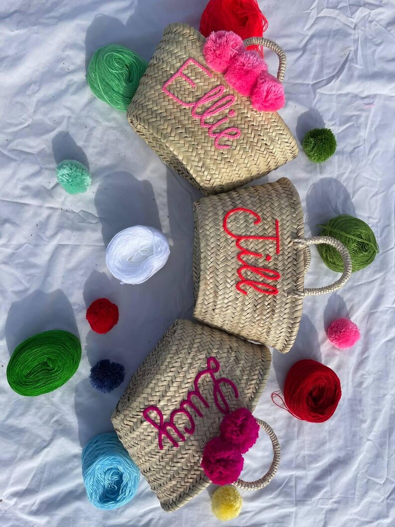 May include: Three straw tote bags with pom poms and personalized names. The bags are Ella,  Tila, and Roma. The bags are surrounded by colorful yarn balls.