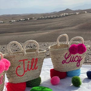 May include: Two straw tote bags with personalized names, "Jill" and "Lucy", in bright pink embroidery. The bags are decorated with colorful yarn pom-poms.