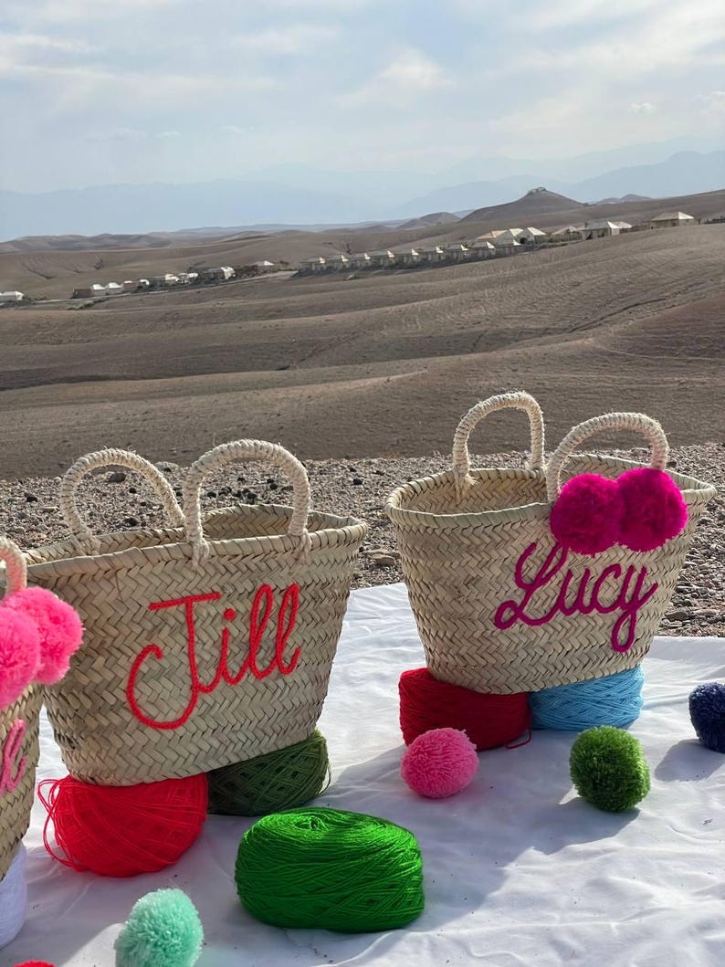 May include: Two straw tote bags with personalized names, "Jill" and "Lucy", in bright pink embroidery. The bags are decorated with colorful yarn pom-poms.
