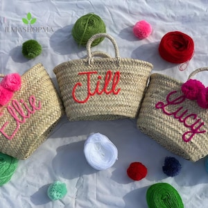Peut inclure: Trois paniers fourre-tout en paille tressée avec des poignées, chacun brodé d'un nom en écriture cursive. Les noms sont "Ellie", "Jill" et "Lucy". Chaque sac a un accent pompon. Des fils et des pompons colorés sont éparpillés autour des sacs.