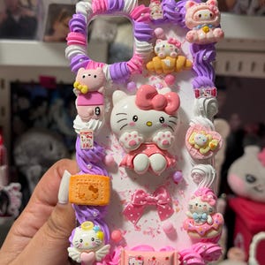 Op de afbeelding: Decoratieve telefoonhoes met een witte basis versierd met diverse roze, paarse en witte versieringen. De hoes bevat meerdere Hello Kitty-beeldjes, strikken en een koekjesvormige bedel. Het ontwerp heeft een getextureerd, gelaagd uiterlijk.