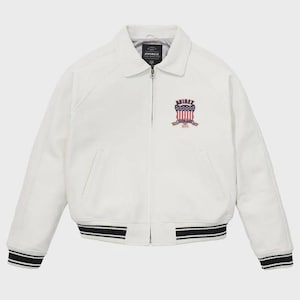 May include: Cream-coloured jacket with a collared neckline and a zip closure. The jacket features a patch with the text "AVIREX" and an American flag design. The cuffs and hem have black and white striped detailing.