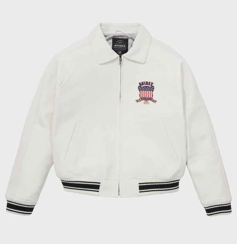 May include: Cream-coloured jacket with a collared neckline and a zip closure. The jacket features a patch with the text "AVIREX" and an American flag design. The cuffs and hem have black and white striped detailing.