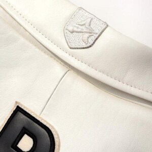 May include: Close-up of a white leather jacket with a stitched design. The jacket features a small, embroidered patch with a stylized bird emblem and a black and white letter "D" patch.