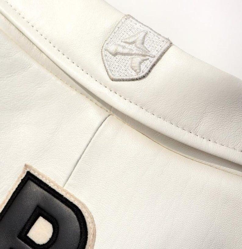 May include: Close-up of a white leather jacket with a stitched design. The jacket features a small, embroidered patch with a stylized bird emblem and a black and white letter "D" patch.