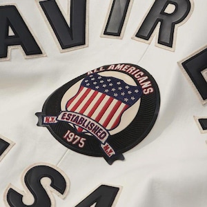 May include: Close-up of a white jacket with black lettering and patches. The main patch features a shield with the American flag and the text "ALL AMERICANS ESTABLISHED 1975 N.Y."