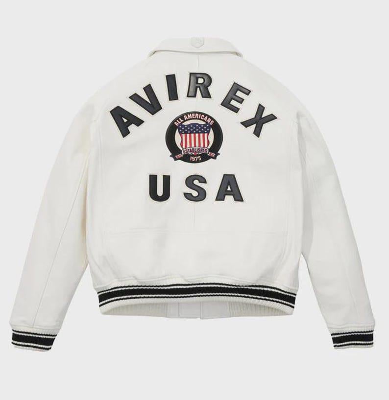 May include: A cream-coloured jacket with black and white striped cuffs and collar. The back displays the word "AVIREX" above a circular emblem with the American flag and the word "USA" below.