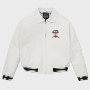 May include: Cream-coloured jacket with a collared neckline and a zip closure. The jacket features a patch with the text "AVIREX" and an American flag design. The cuffs and hem have black and white striped detailing.
