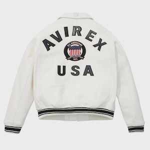 May include: A cream-coloured jacket with black and white striped cuffs and collar. The back displays the word "AVIREX" above a circular emblem with the American flag and the word "USA" below.