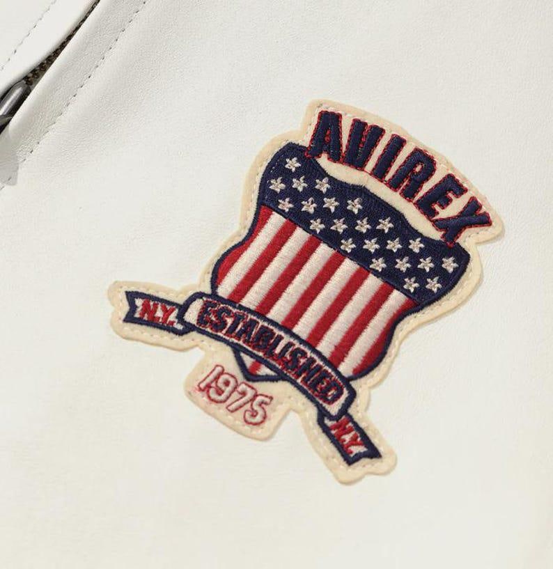 May include: A white leather jacket with a patch displaying the word "AVIREX" in red, a blue field of stars, and red and white stripes. The patch also includes the text "N.Y. ESTABLISHED 1975."