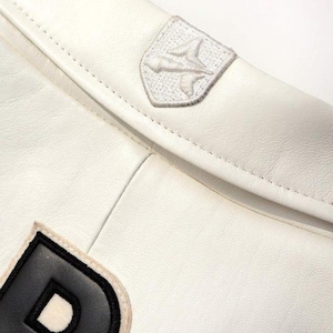 May include: Close-up of a white leather jacket with a stitched design. The jacket features a small, embroidered patch with a stylized bird emblem and a black and white letter "D" patch.