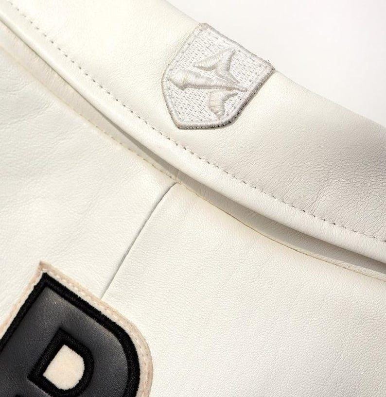 May include: Close-up of a white leather jacket with a stitched design. The jacket features a small, embroidered patch with a stylized bird emblem and a black and white letter "D" patch.
