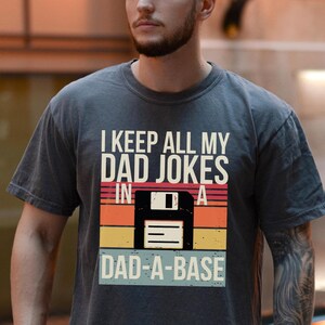 Könnte beinhalten: Ein dunkelgraues T-Shirt mit einer Retro-Grafik, auf der "I keep all my dad jokes in a dad-a-base" steht. Die Grafik zeigt eine Diskette mit einem Sonnenuntergangs-Hintergrund.