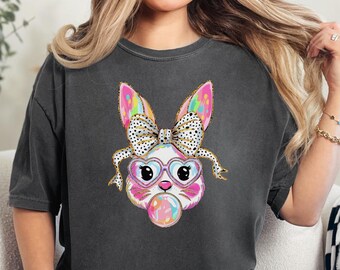 Peut inclure: Un t-shirt gris anthracite avec un lapin de dessin animé coloré portant des lunettes de soleil roses et un nœud. Le lapin souffle une bulle.