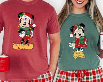 Puede incluir: Dos camisetas a juego con temática navideña, una burdeos y otra verde, cada una con un personaje de dibujos animados de Mickey Mouse envuelto en luces navideñas. Ambas se combinan con pantalones de pijama de cuadros rojos. Una persona sostiene una taza roja.