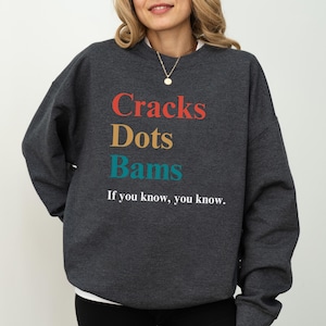 May include: Dark gray sweatshirt with the words "Cracks Dots Bams" in red, gold, and teal. Below the words is the phrase "If you know, you know." The sweatshirt is a crew neck style.
