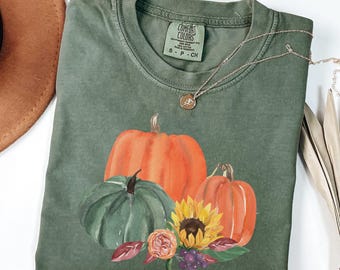 May include: Green Comfort Colors t-shirt with a watercolour design of pumpkins, a sunflower, and autumn leaves. A silver necklace with a pendant rests on the shirt. The shirt has a small tag that says "S - P - CH".