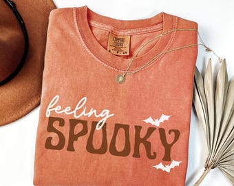 May include: An orange Comfort Colors t-shirt with the words "feeling SPOOKY" in white and brown, with two white bat graphics. A gold necklace with a pendant is draped across the shirt. A brown hat and dried palm leaf are in the background.