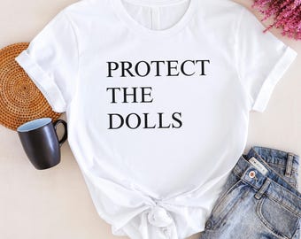May include: White t-shirt with the text "PROTECT THE DOLLS" in black. The shirt is knotted at the waist and paired with blue denim jeans. A black mug and woven basket are visible on the left.