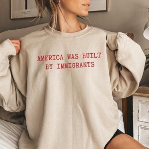May include: A beige sweatshirt with the text "America was built by immigrants" in red.