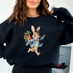 May include: Black sweatshirt featuring a cartoon image of a rabbit wearing a blue jacket and carrying a basket of carrots.