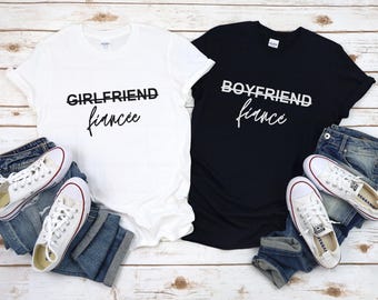 May include: Two white t-shirts with black text. One shirt says "Girlfriend fiance" and the other says "Boyfriend fiance".