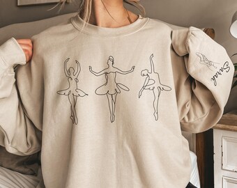 Könnte beinhalten: Beiges Sweatshirt mit einer schwarzen Strichzeichnung von drei Ballerinas. Der rechte Ärmel hat eine Skizze einer Ballerina und den Namen "Sarah" in Schreibschrift. Das Design ist in schwarzer Tinte.