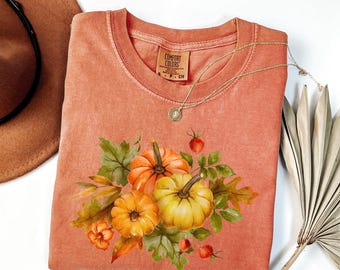 May include: A coral-coloured Comfort Colors t-shirt with a watercolour design of pumpkins, leaves, and berries. The shirt has a round neckline and short sleeves. A gold necklace with a pendant is draped across the shirt. A brown hat and dried palm leaf are in the background.