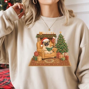 May include: A sand-coloured crewneck sweatshirt featuring a graphic of a pug wearing a Santa hat, sitting in a tan armchair reading a book. The scene includes a decorated Christmas tree, a fireplace with a stocking, and wrapped gift boxes.