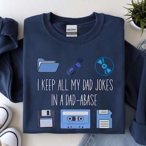 Può includere: Felpa blu navy con la scritta "I KEEP ALL MY DAD JOKES IN A DAD-ABASE" e grafiche di una cartella, una chiavetta USB, un CD, un floppy disk, una musicassetta e un altro floppy disk. La felpa è accanto a un paio di scarpe da ginnastica e jeans.
