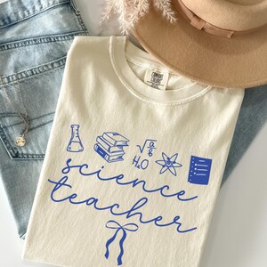 Può includere: T-shirt color crema con grafica blu a tema scientifico e la scritta "science teacher". La maglietta è abbinata a jeans chiari, un cappello marrone e una collana d'argento.