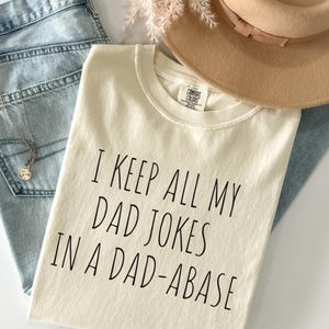 Può includere: T-shirt color crema con la scritta "I KEEP ALL MY DAD JOKES IN A DAD-ABASE" in nero. La maglietta è appoggiata su jeans azzurri, una collana dorata e un cappello beige.