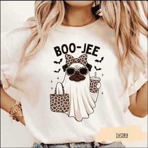 May include: An ivory t-shirt featuring a graphic of a pug dressed as a ghost, wearing leopard print sunglasses and a matching bow. The pug holds a leopard print tote bag and a drink cup. The text BOO-JEE is printed above with small black bats.