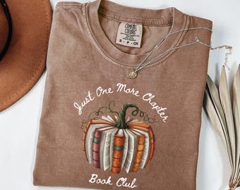 May include: A brown Comfort Colors t-shirt with the text "Just One More Chapter Book Club" and a pumpkin design made of books. A silver necklace with a pendant and a brown hat are also visible.