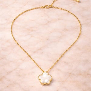 May include: A gold-colored necklace with a white flower-shaped pendant. The pendant has a gold-colored border. The necklace chain is delicate and adjustable. The background is a light pink marble pattern.