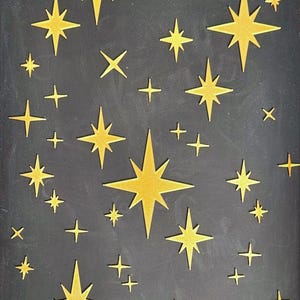 May include: A rectangular black chalkboard with a white distressed wooden frame. The chalkboard is decorated with various sizes of gold stars and crosses. The stars are in different sizes and shapes, creating a celestial theme.