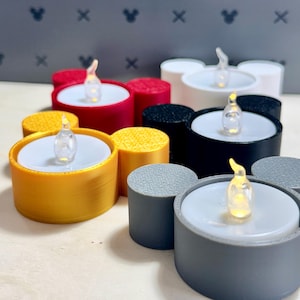 May include: A collection of five circular tea light candle holders with two smaller circular side attachments, resembling a mouse silhouette. The holders are in red, yellow, white, black, and grey, each containing a white tea light with a glowing flame.