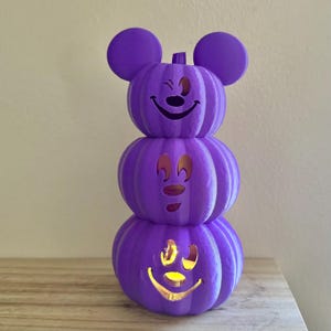 May include: A vertical stack of three purple pumpkins featuring Mickey Mouse-inspired ears on the top pumpkin. Each pumpkin has a carved face with cutouts, and the bottom pumpkin is illuminated from within, casting a warm yellow glow through the eyes and mouth.