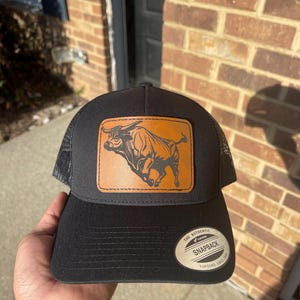 May include: Black trucker hat with a brown leather patch featuring a black silhouette of a bull. The hat has a black mesh back and a silver metal label that reads "The Authentic Snapback Yupoong Since 1974."