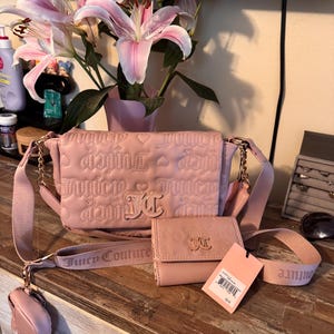 May include: A pink Juicy Couture handbag, wallet, and coin purse set. The handbag has a quilted design with the brand name and logo. The wallet and coin purse match the handbag. The set is displayed on a wooden surface with a pink strap and a tag.