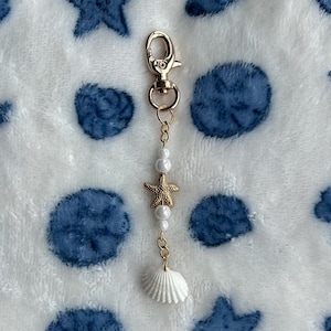 Pode incluir: Um chaveiro com um pingente de ouro em forma de concha branca, uma estrela do mar dourada e contas de pérolas brancas. O chaveiro está sobre um fundo branco com desenhos de estrelas do mar e conchas azuis.