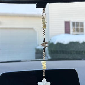 May include: A white flower charm hangs from a gold chain with yellow and brown beads. The charm is attached to a rearview mirror in a car.