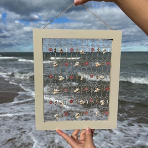 May include: A white wooden frame with a wire mesh background. The mesh is decorated with small pink and white beads, seashells, and gold beads. The frame is hanging from a chain.